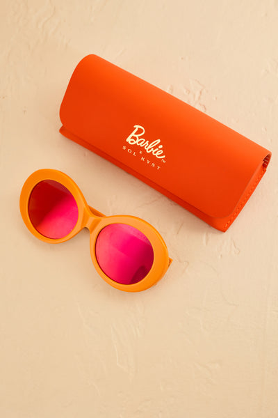 Barbie Sol Kyst round orange sunglasses with pink-tinted lenses, displayed alongside an orange protective case on a textured beige surface.