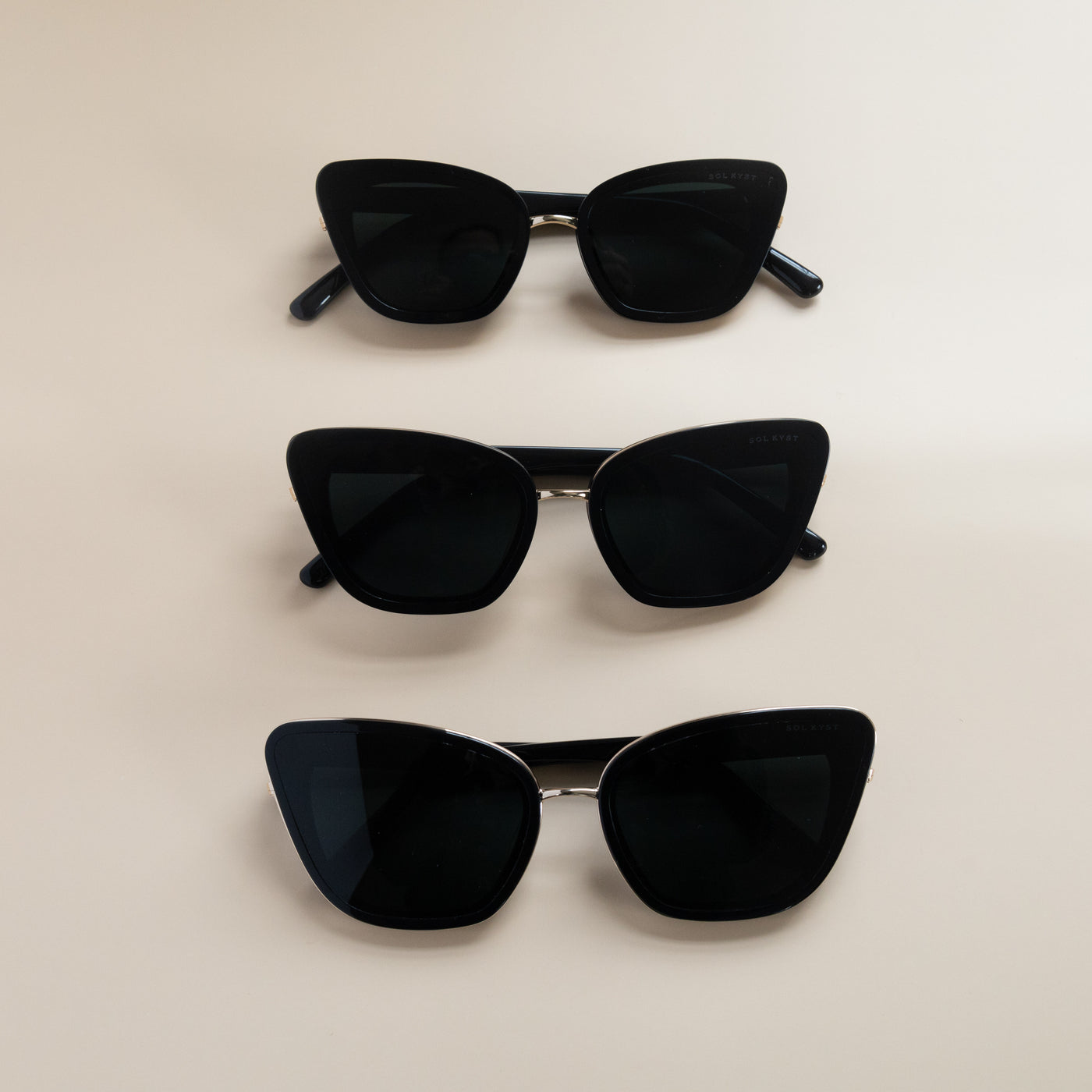 Three pairs of Sol Kyst women's cat eye sunglasses, with black frames and dark tinted lenses, arranged vertically on a beige surface.