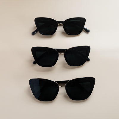 Three pairs of Sol Kyst women's cat eye sunglasses, with black frames and dark tinted lenses, arranged vertically on a beige surface.
