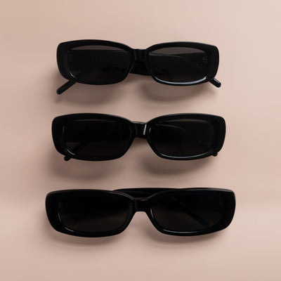 Three pairs of rectangular black sunglasses with dark tinted lenses arranged vertically on a beige surface.