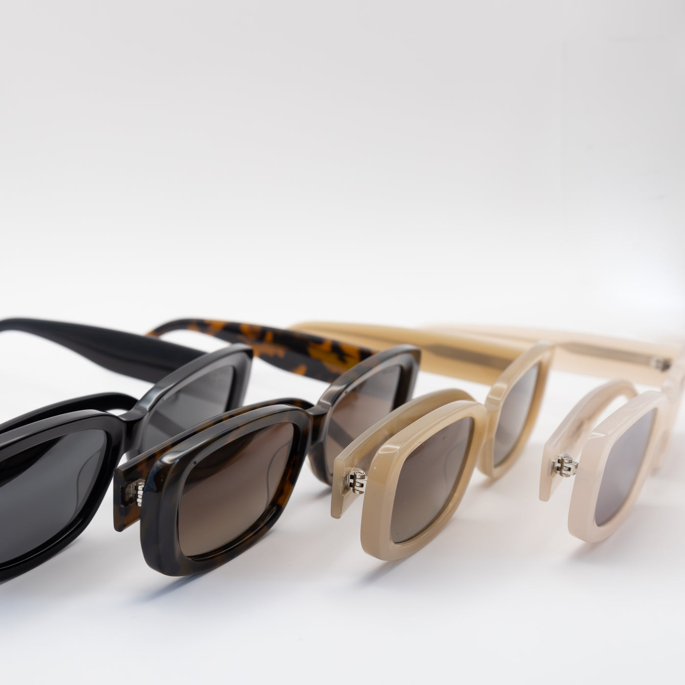 Four rectangular sunglasses in black, tortoise, beige, and cream frames with tinted lenses, displayed in a row at an angle on a white surface.