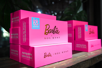 Pink with gold accents "Barbie x SOL KYST" product boxes stacked on a windowsill.