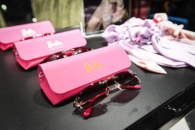 Pink Barbie sunglasses next to their signature pink cases, accompanied by pastel accessories in the background.